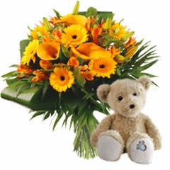 Mélange de fleurs aux couleurs chaudes, tiges moyennes rehaussées d'une verdure éclatante accompagné d'un ours brun en peluche, réalisé avec des matiè